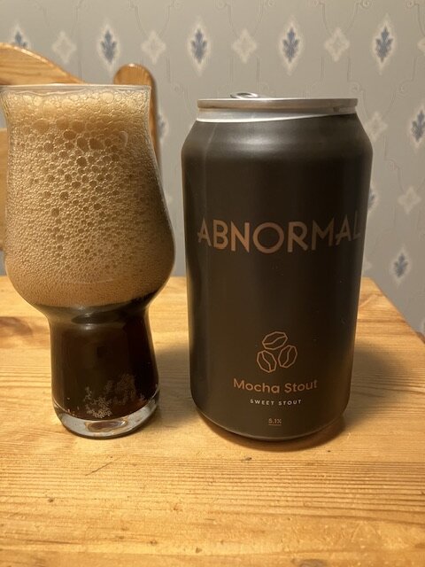 Öl med rikligt skum i glas bredvid burk märkt "ABNORMAL Mocha Stout Sweet Stout".