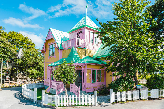 Färgglatt, pastellfärgat hus med spiraltorn, vita staket, lekplats i bakgrunden, solig dag, frodig grönska.