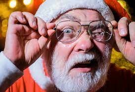 Man förklädd till jultomten ser förvånad ut, lyfter glasögonen, bär tomtemössa, oskarp röd bakgrund.