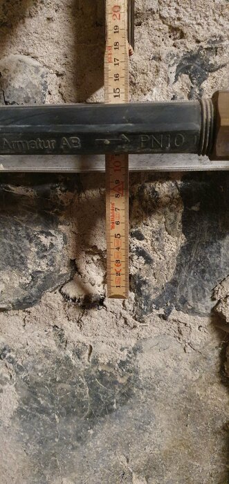 Måttband bredvid rör och betongvägg, antagligen för att mäta djup eller avstånd.