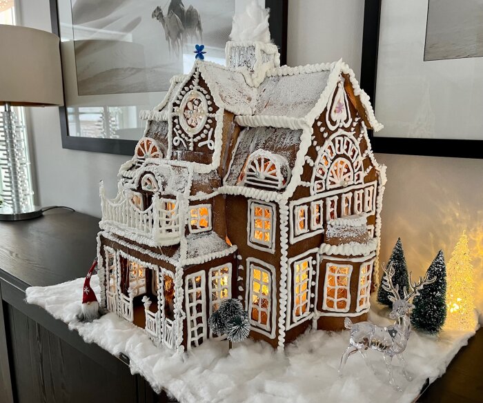 Detaljerat pepparkakshus med belysning, snöeffekt och juldekorationer på en bordsyta inomhus.