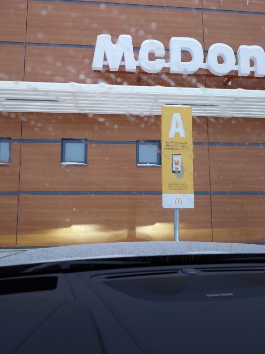 Snötäckt bil framför McDonald's med beställningsstolpe, skylt A, vintern, kvällsbelysning speglar sig på fasaden.