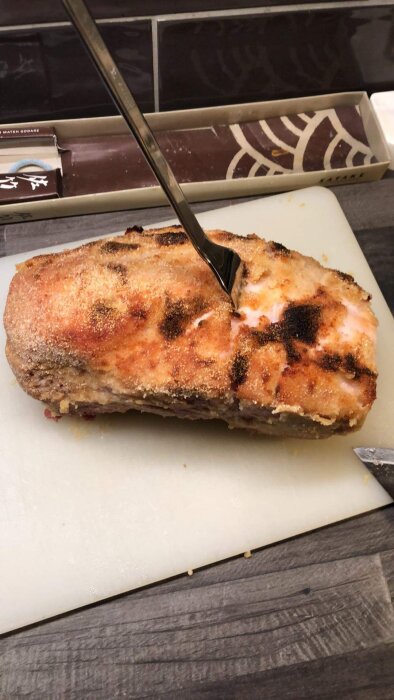 Grillad eller stekt mat, kanske kött, med brända delar, på skärbräda med gaffel, köksmiljö.