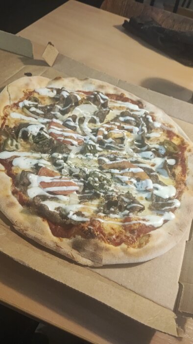 Pizza med olika pålägg i kartong, sannolikt takeaway, med vit såsdrizzle överst.