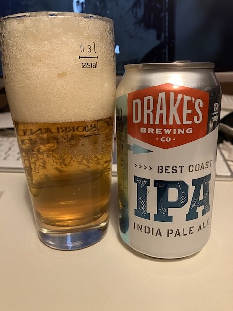 Ett glas öl bredvid en ölburk märkt "Drake's Brewing IPA" på ett skrivbord.