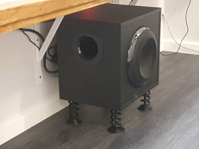 Svart subwoofer på fjädrande fötter i ett hörn, bredvid vit vägg, träarbete ovanför, kablar synliga.