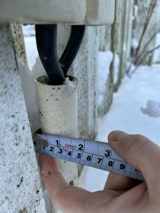 Hand håller måttband vid pelare med kablar och snö, utomhus, dagtid, mäter avstånd.