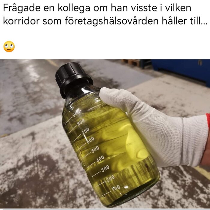 Hand i vante håller plastflaska med gul vätska, industriell bakgrund, mätmarkeringar på flaskan.