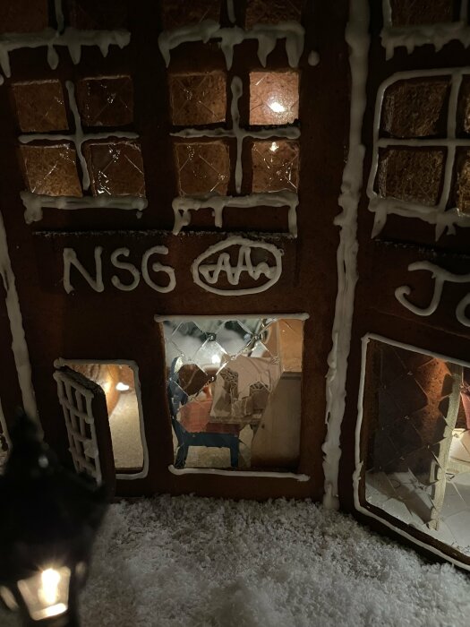 Pepparkakshus med ljus inuti, snölik dekoration, fönster, "NSG" skrivet, mörk bakgrund.