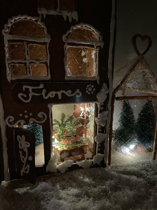 Ett pepparkakshus med belysning, snöeffekt, texten "Flores" och dekorativa detaljer; närbild, mysig, julstämning.