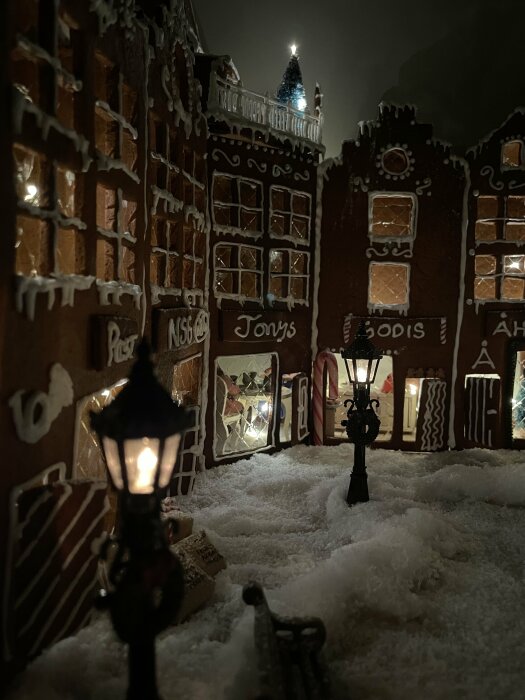 En nattlig scen av pepparkakshus med snö, gatlyktor, fönster och textdekorationer, ser ut som en miniatyrstad.