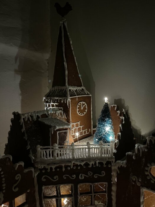 Pepparkakshus med torn, klocka, dekorationer och gran, upplyst i dunkel belysning med skuggor på väggen.