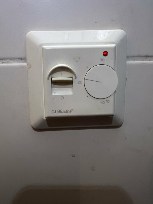 En smutsig termostat med vred och på-av-brytare på en vägg, röd indikatorlampa tänd, märke "OJ Microline".