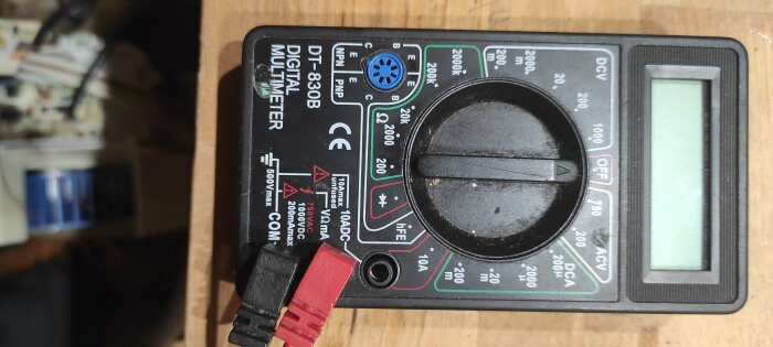 Digital multimeter lutad neråt, svart selektorknapp, röd och svart testledning; bakgrund med elektriska komponenter.