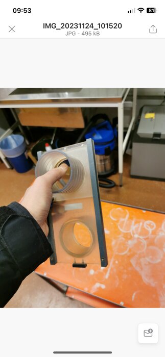 Hand håller en genomskinlig akrylplatta med urklippta cirklar och metallskruvar i ett verkstadsutrymme.