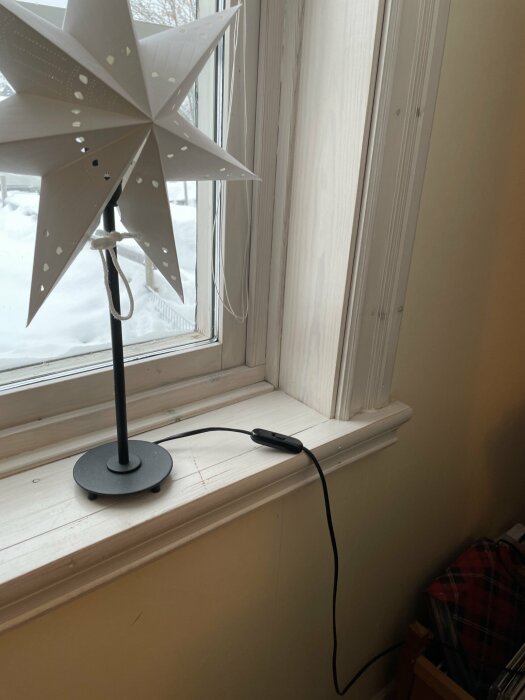 Fönsterstjärna på fot, fönsterkarm, vitmålat trä, snökant synlig utomhus, kabel hänger ner, grå vägg.