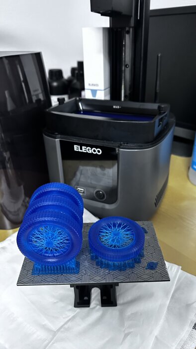 Två blå, nyligen 3D-utskrivna objekt, med en SLA 3D-skrivare av märket ELEGOO i bakgrunden.