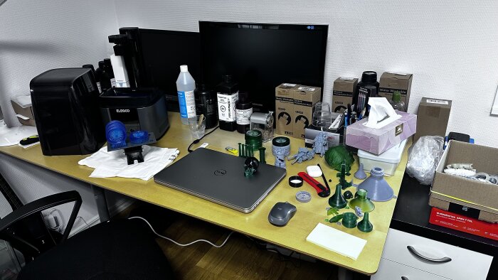 Arbetsbord med 3D-skrivare, dator, förbrukningsmaterial och olika 3D-utskrivna objekt. Rörigt och teknikinriktat.