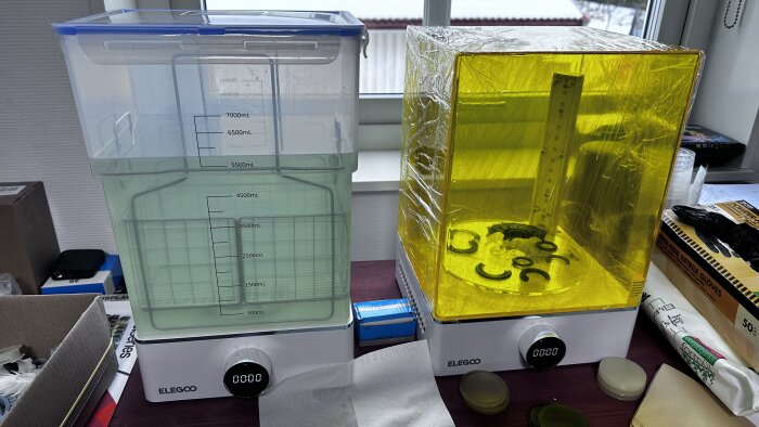 Två Elegoo tvätt- och härdningsstationer för 3D-utskrifter, en tom, en innehåller gul resin. Arbetsmiljö.