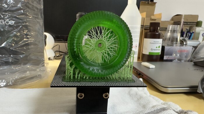 Ett grönt plasthjul från en 3D-skrivare på arbetsbänk, med stödstrukturer kvar, framför oordnad skrivbordsbakgrund.