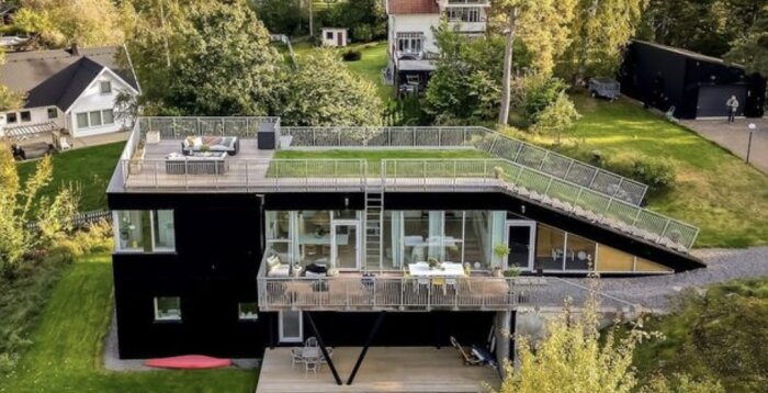 Modernt svart hus med stora fönster, takterrass, omgivet av grönska i ett villaområde.