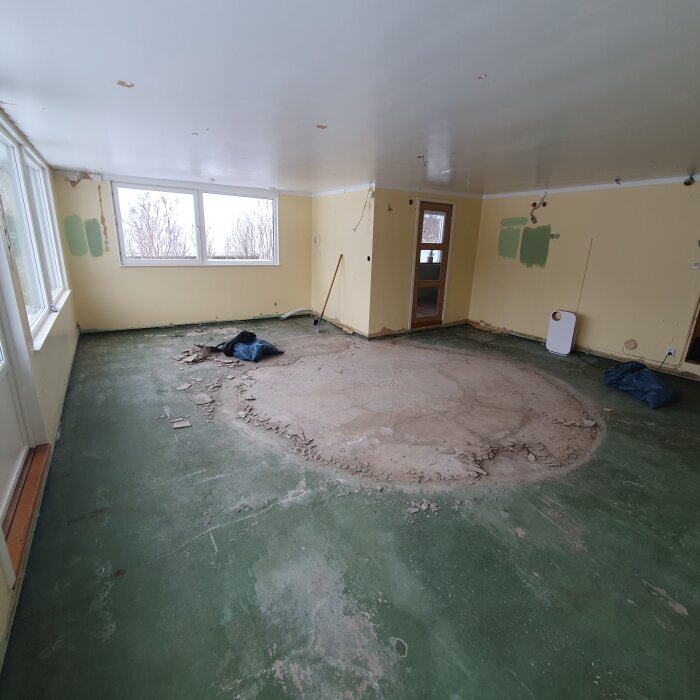 Tomt rum under renovering, utsatt betonggolv, väggar med avlägsen tapet, fönster, skräp på golvet.