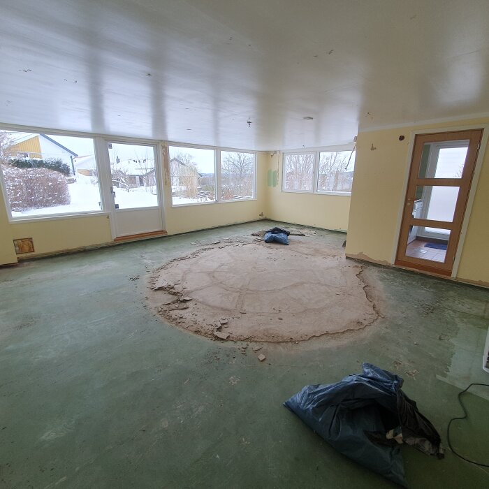 Ett tomt rum under renovering med utsikt över vintern, borttagen golvbeläggning, och byggmaterial på golvet.