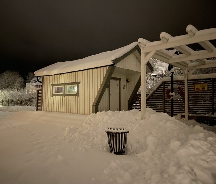 Hus täckt av snö på natten, upplyst fönster, snötäckt papperskorg, trätrappa, snöig omgivning.