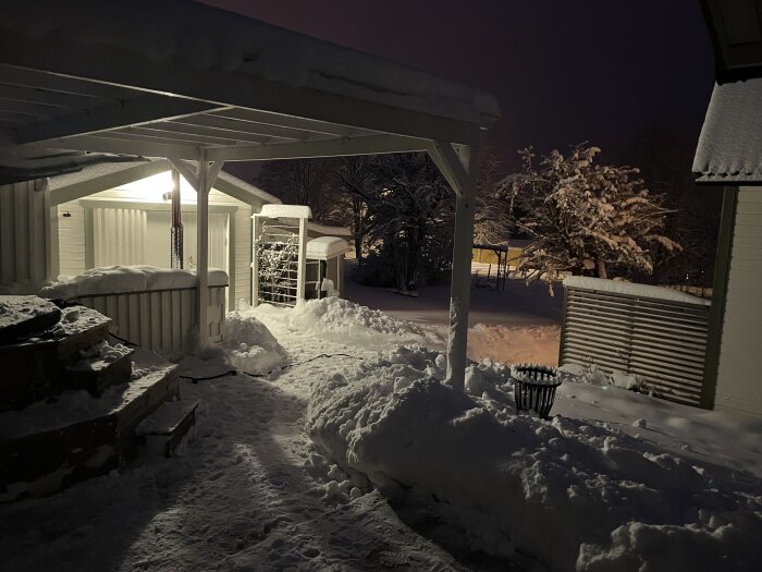 Nattbild av en snöig trädgård med carport, skjul, och snötäckta träden under belysning.