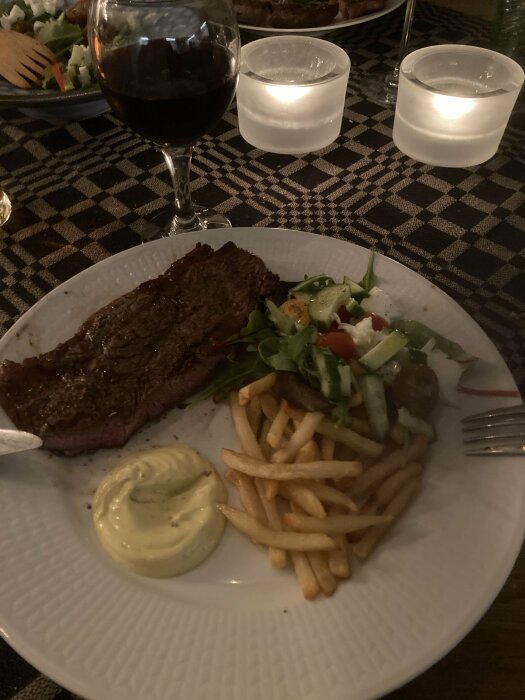 En middagstillfälle med biff, pommes frites, sallad, sås och rödvin. Ljus och mönstrad duk i bakgrunden.