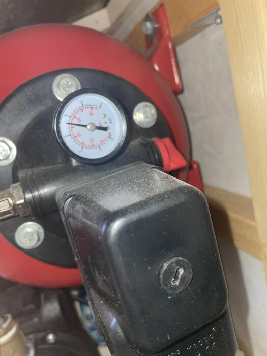 Röd trycktank med manometer, svart plasthus i förgrunden, monterad på trästruktur. Industriell eller verkstadsutrustning.