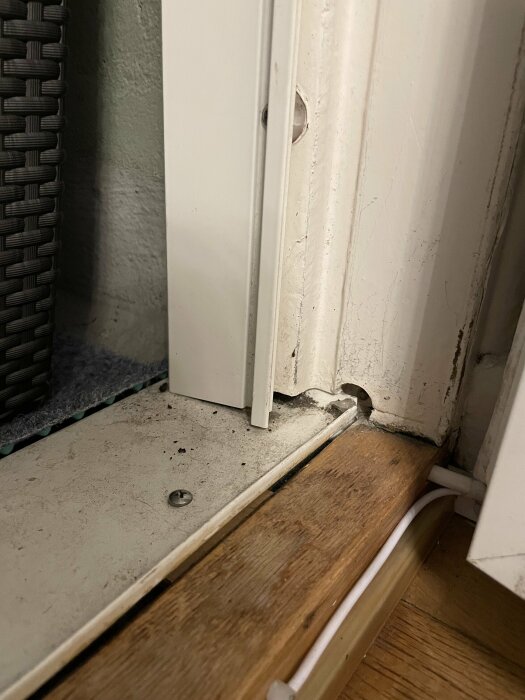 Närbild av ett hörn, smutsigt golv, kablar, vit dörr med sliten kant och en grå vägg.