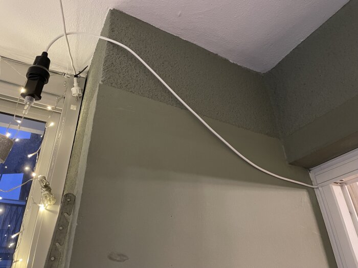 Hörn med belysning: glödlampa i sladd, ljusslinga, vit kabel längs vägg och tak.