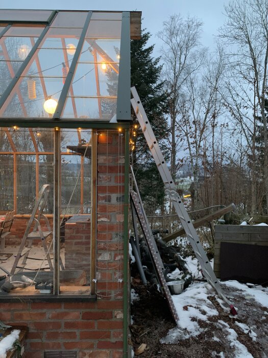 Ett växthus med tegelgrund, upplyst av ljusslingor, mot vinterbakgrund vid skymningen, med stege lutad mot det.