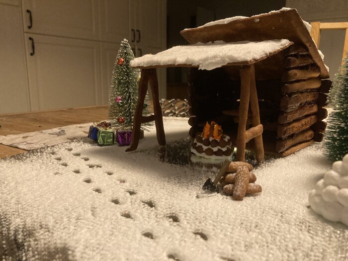 Ett pepparkakshus täckt av florsocker "snö" med miniatyrjulgran, presenter och tårta på bordet.