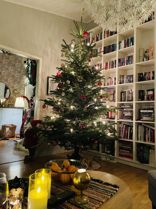 Julgran med röda kulor och ljusslingor inomhus, bokhylla, dekorationer, mysig stämning.