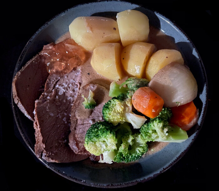 Mörk tallrik med roastbiff, potatis, morötter, broccoli och sås, mörk bakgrund.