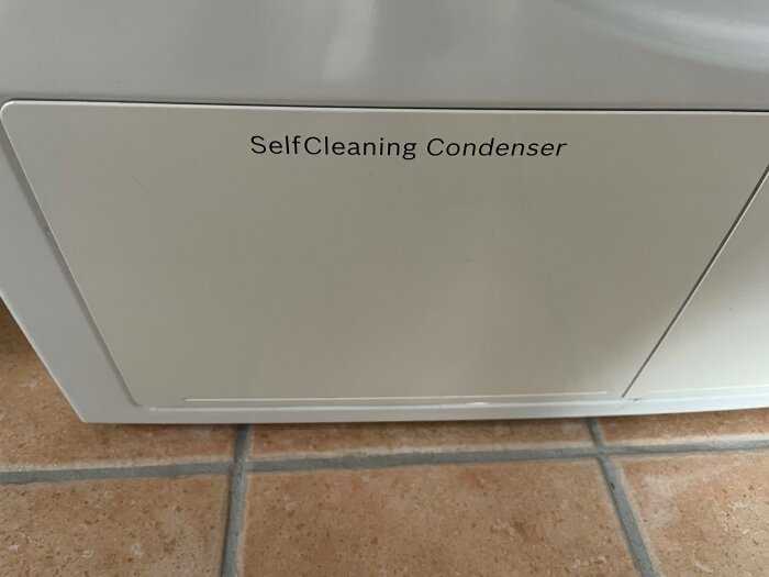 Vit apparat med text "SelfCleaning Condenser", troligtvis en del av en torktumlare, på kakelgolv.