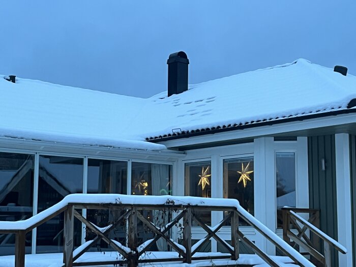 Ett snötäckt hus i skymningen med stjärnformade ljus i fönstren och en veranda.