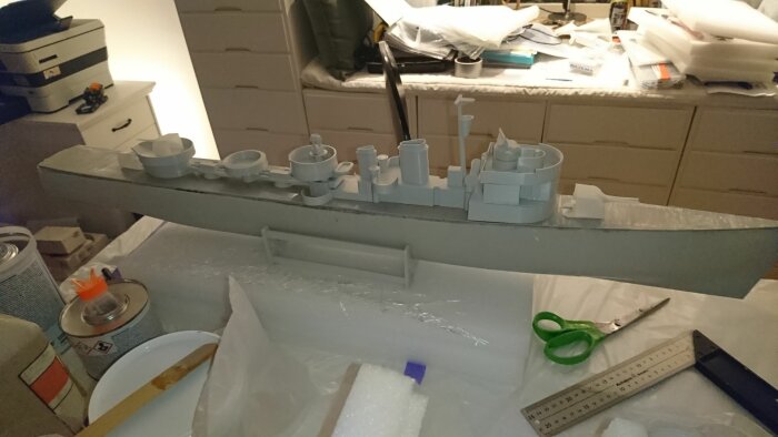 Ett modellskepp under konstruktion omgivet av verktyg och material på ett rörigt arbetsbord.