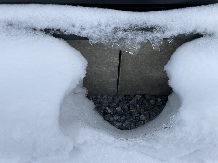 Struktur i metall täckt av snö och is med småstenar synliga nedanför, vinterkyla, utomhus, dagsljus.