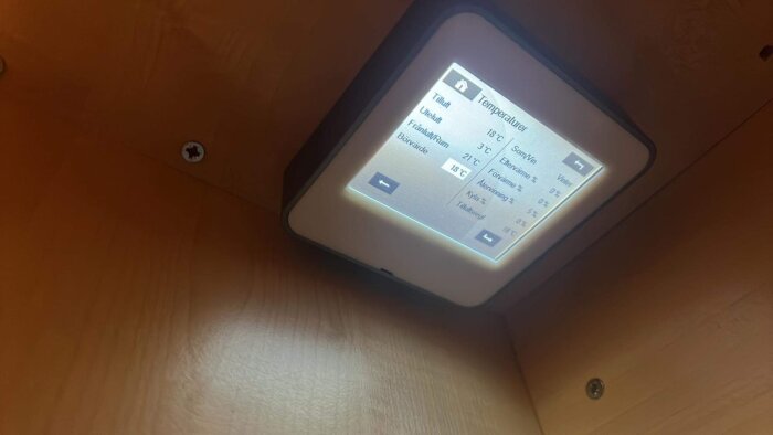 Digital termostat visar inomhustemperaturer på svenska, monterad på träpanel, sned fotovinkel, belysning från skärmen.