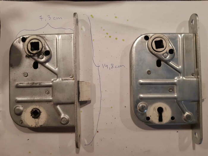 Två metalliska dörrlåsdelar ligger på ett papper med måttangivelser, visar fram- och baksida, använda och nya.