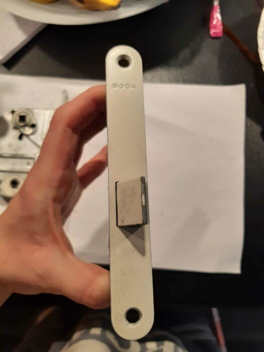 Hand håller en vit metallskena märkt "BODA" ovanför suddig bakgrund med papper och föremål.