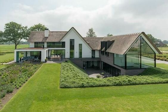 Modernt hus med stora fönster, grön trädgård, asymmetriskt tak, öppen terrass, landsbygdsmiljö.