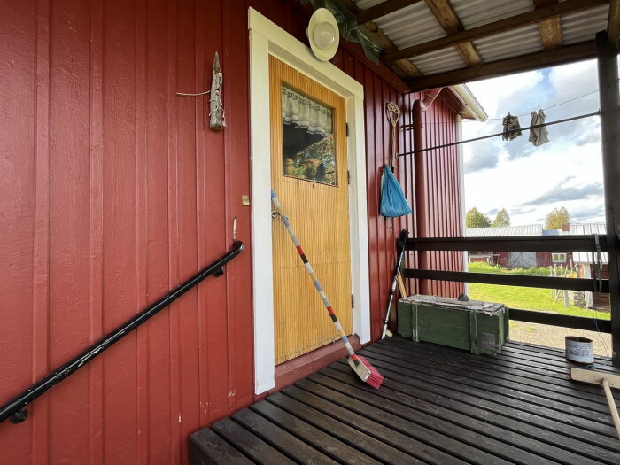 Träveranda, röd vägg, dörr, kvast, soppåse, målarburk, gröna fält, molnig himmel.