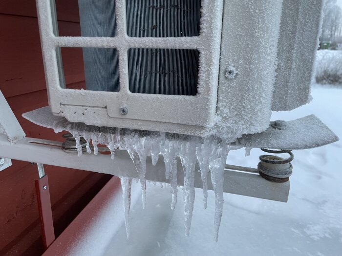 Luftvärmepump täckt av frost och istappar, vinterkyla, utomhus, röd vägg i bakgrunden.