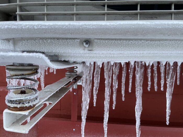 Vitt frosttäcke och is istappar på metallstruktur och gångjärn, kallt väderfenomen, röd bakgrund.