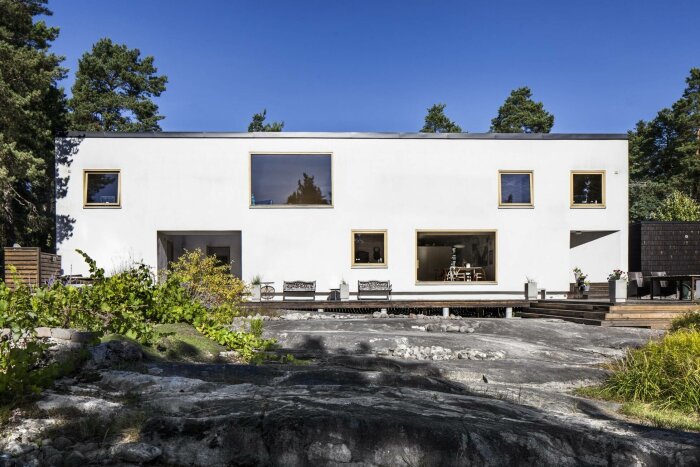 Vitt modern hus med oregelbundna fönster, terrass, klippor och skogsbakgrund.