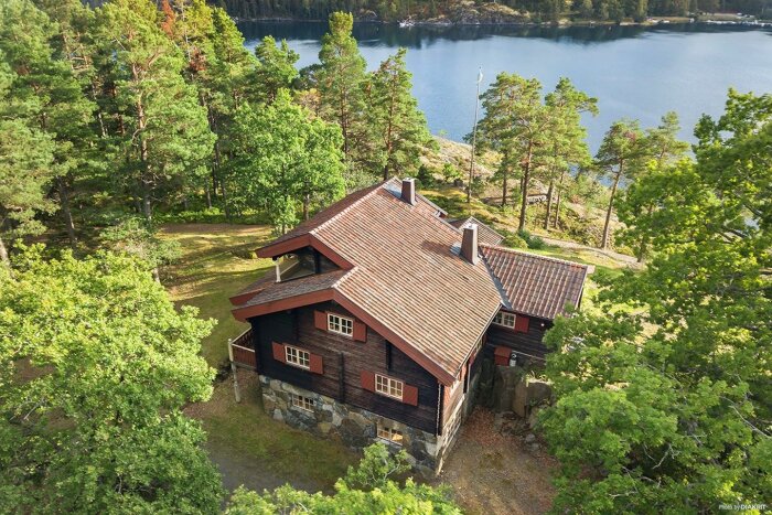 Röd stuga i skog nära sjö, tomt, natur, lugnt, takpannor, veranda, stenfundament, djupgrön skogsmark, avskildhet.